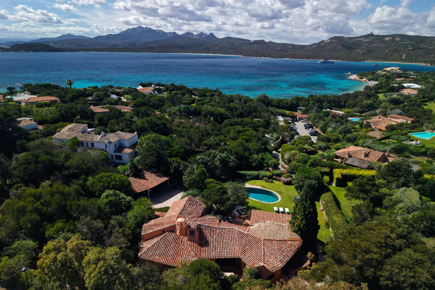 Aerial view of a Mediterranean villa with a terracotta roof, pool, and lush gardens overlooking a turquoise bay.