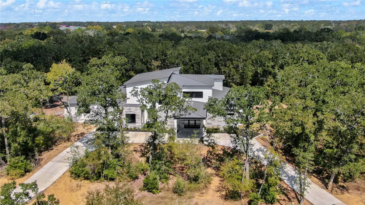 Aerial view of a modern white house with a gray roof, surrounded by green trees and a long driveway.