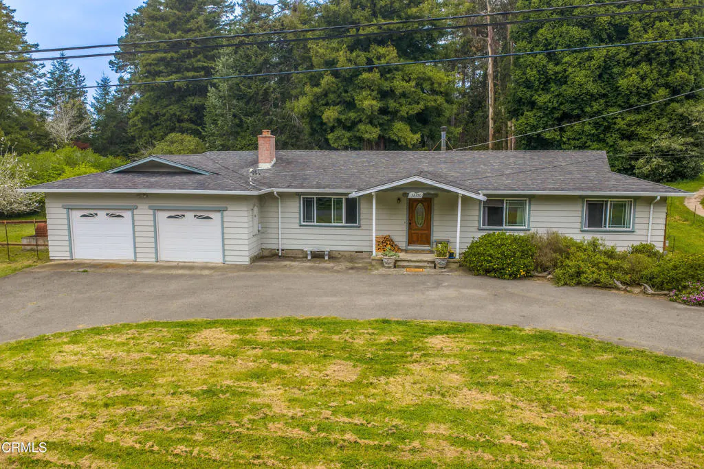 A single-story, light-gray house with a gray roof, a brick chimney, and a two-car garage. The house has a brown front door and is surrounded by green trees.