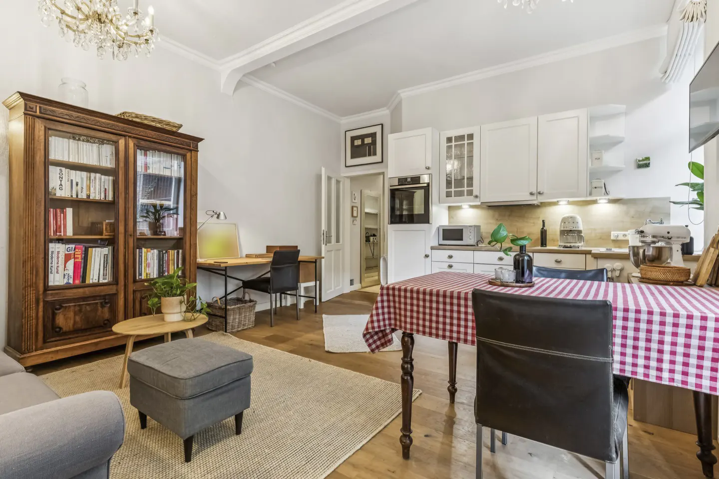 Open-plan living area with a wooden bookcase, a small desk, and a dining table with a red-and-white checkered tablecloth.
