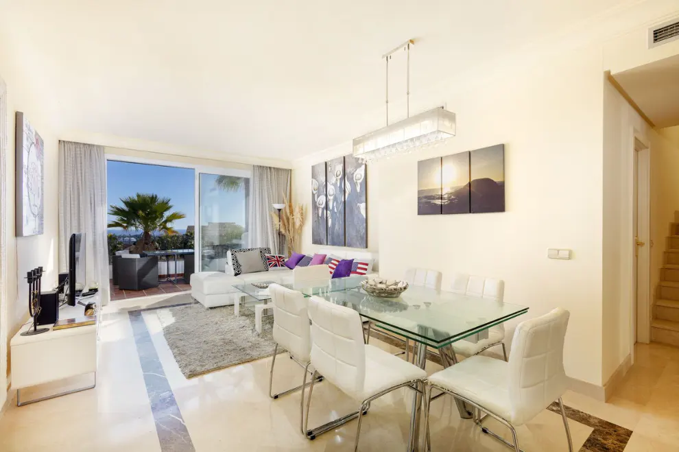Bright, open-plan living and dining area with glass table, white chairs, and sofa. Balcony with palm tree view. Modern art on the wall.