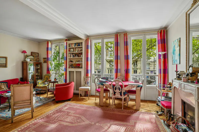 Bright living room with hardwood floors, a pink rug, and large windows with colorful curtains. A dining table and chairs sit near the windows.