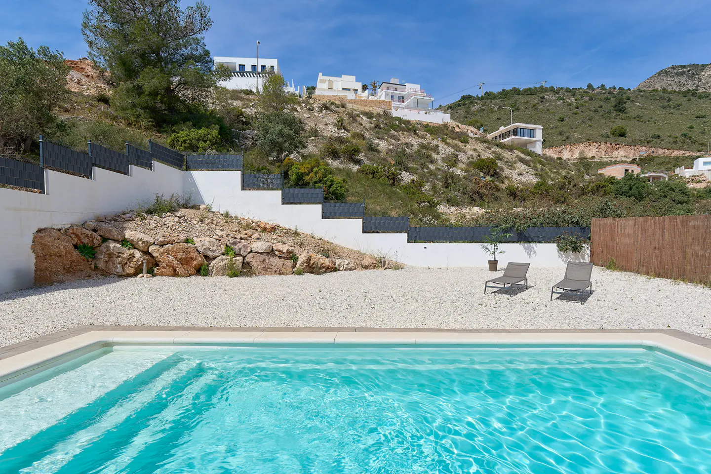 A bright blue swimming pool with steps, surrounded by white gravel and two lounge chairs. Houses are visible on the hillside in the background.