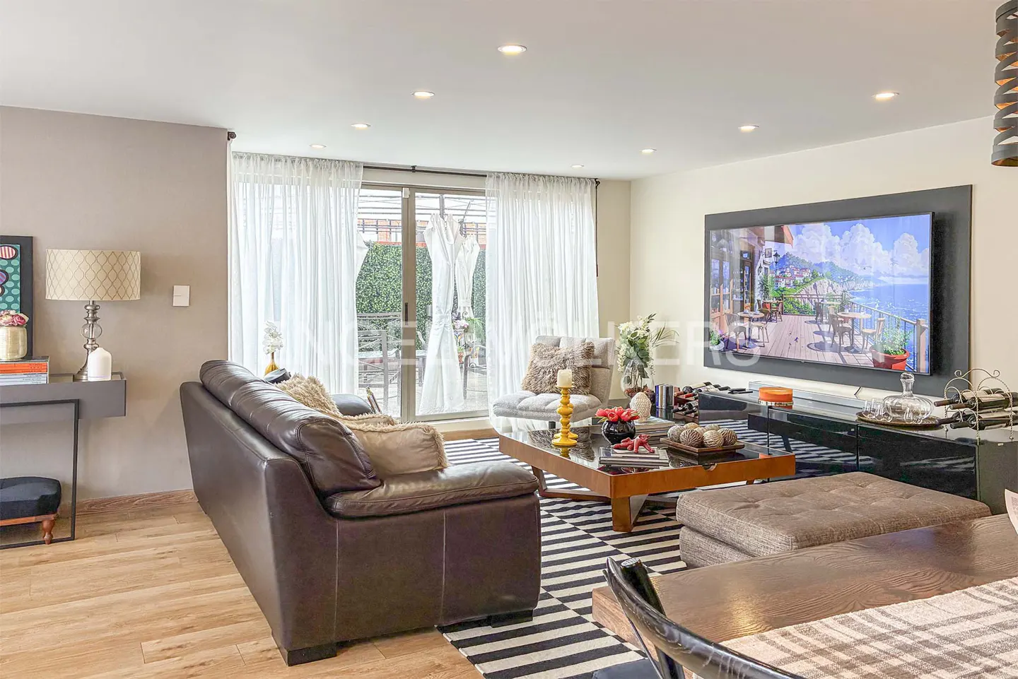 Living room with a brown leather sofa, striped rug, and a TV showing a coastal scene. White curtains frame a glass door to a patio.