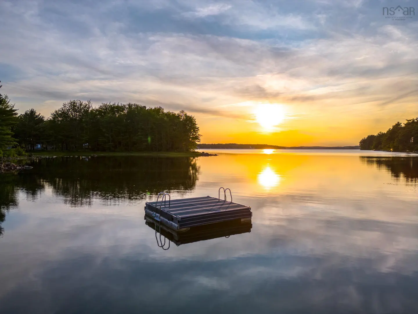 Sunset over a calm lake with a floating dock in the foreground. Trees line the shore.