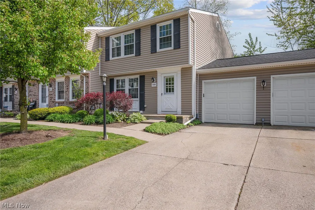 Two-story tan house with black shutters, white door, and two-car garage. Green lawn and trees in the front.