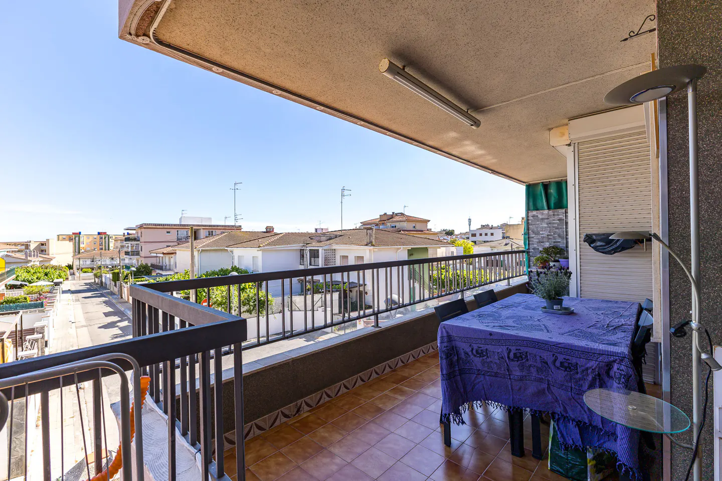 Balcony view with brown tile floor, black railing, and a table covered with a blue cloth. Houses and blue sky are visible in the background.