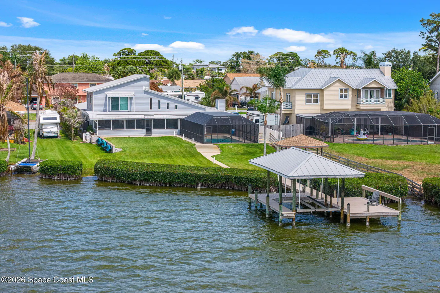 Waterfront homes with docks on a sunny day. One house is gray with a screened-in pool, the other is beige.