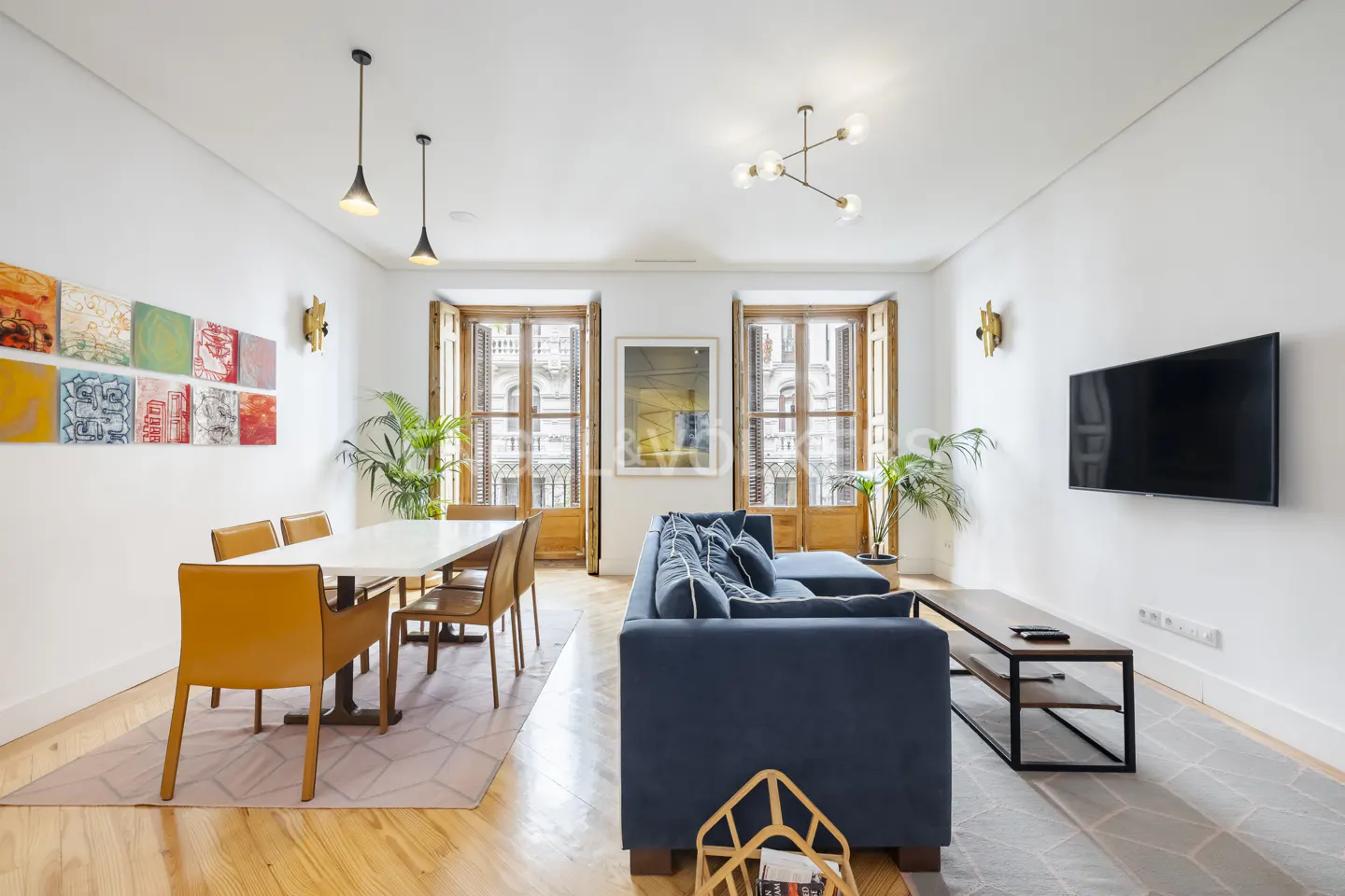 Bright living room with wood floors, white walls, and large windows. A blue sofa faces a TV, and a dining table sits near colorful artwork.
