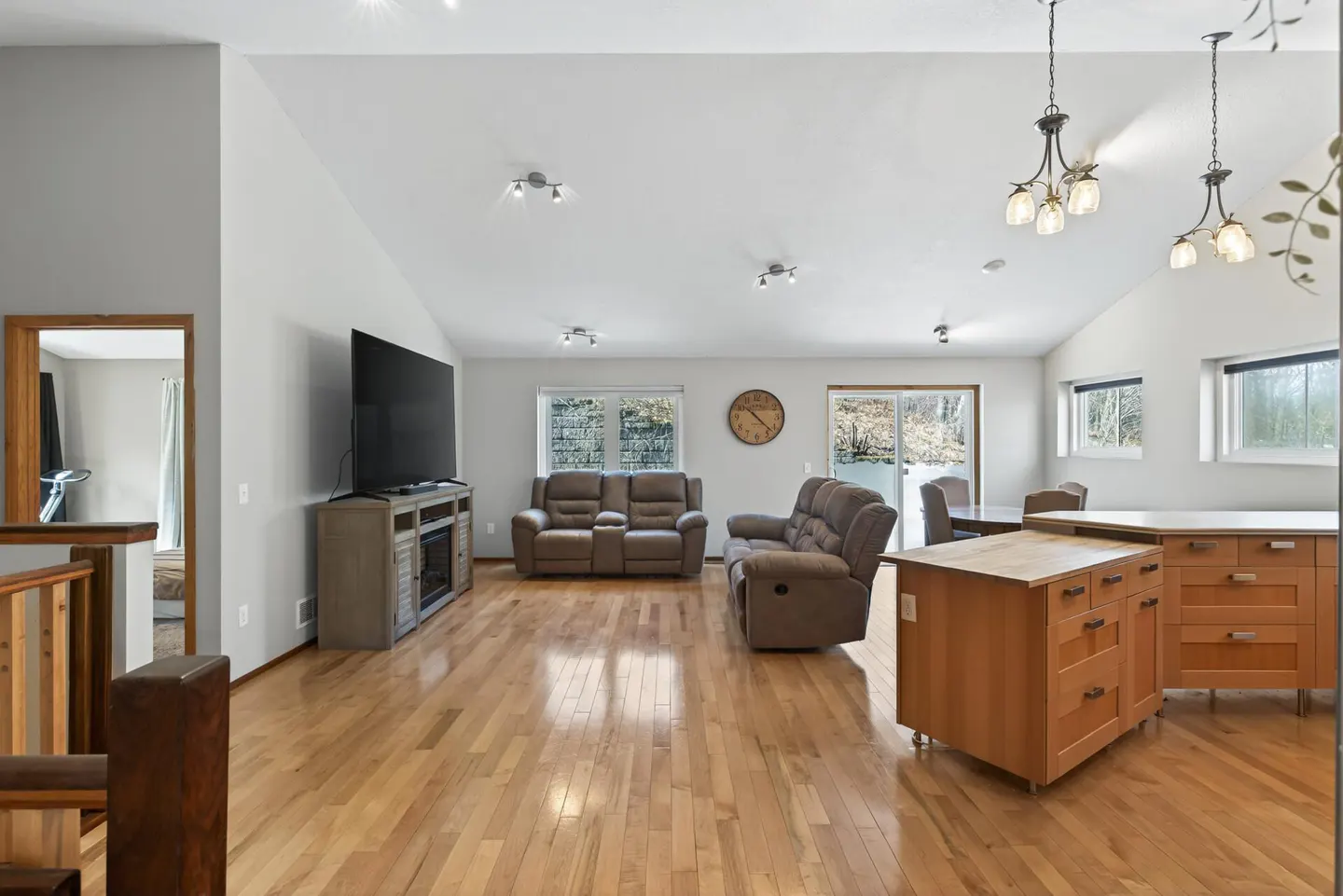 Open-concept living room with hardwood floors, gray walls, and vaulted ceilings. Two brown sofas face a TV on a gray stand. Kitchen island in foreground.