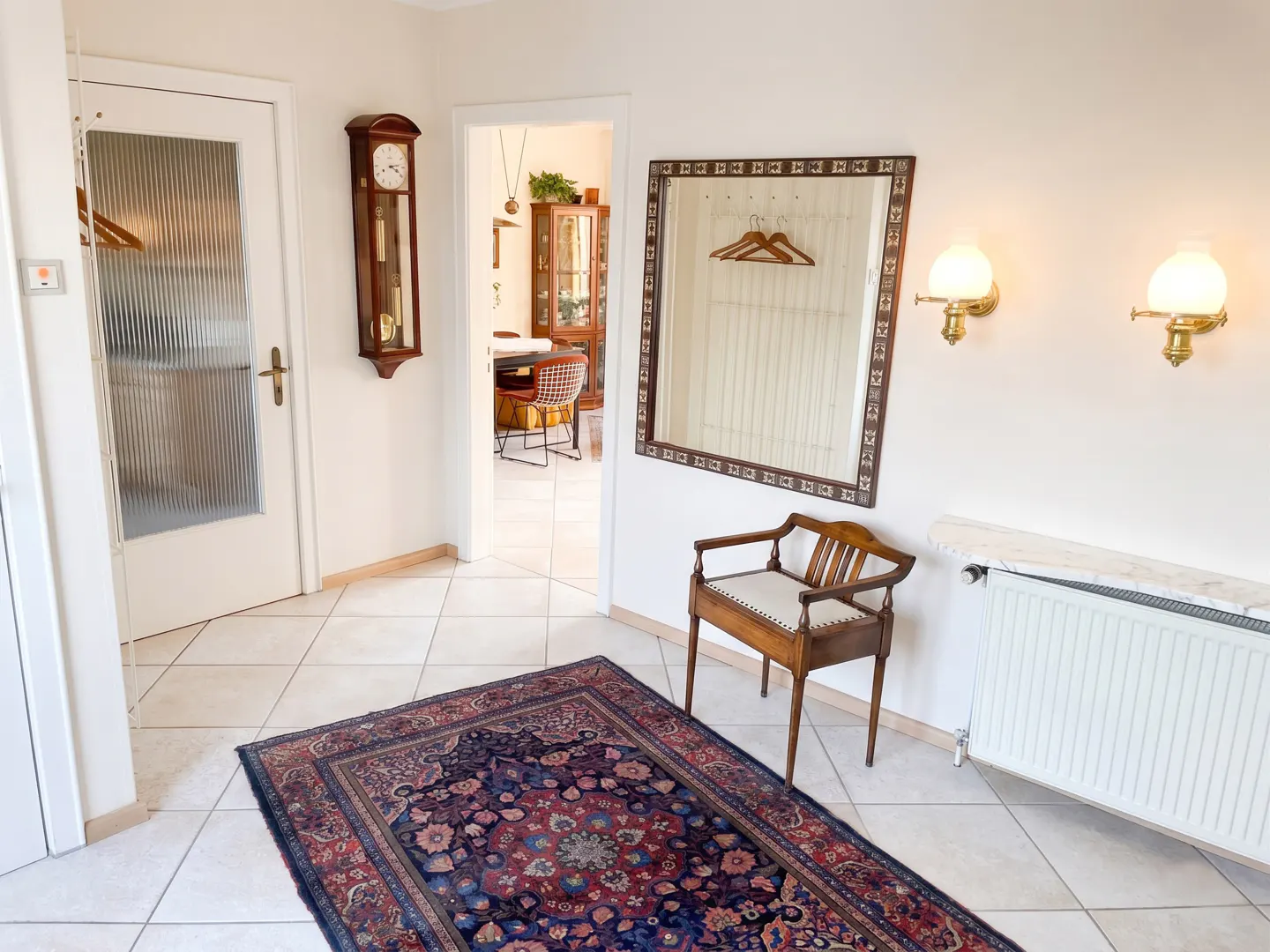 A bright hallway with a mirror, wooden chair, and blue rug on a tiled floor. A grandfather clock hangs on the wall.
