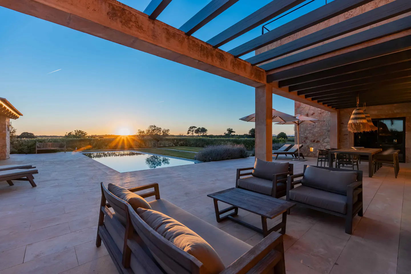 Outdoor patio with lounge seating, dining table, and pool overlooking a field at sunset.