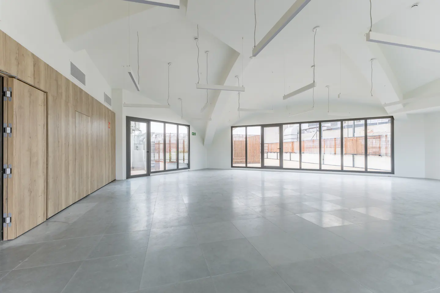 Empty room with gray tile floor, white walls, and a high, peaked ceiling. Large windows and a wood-paneled wall add texture.