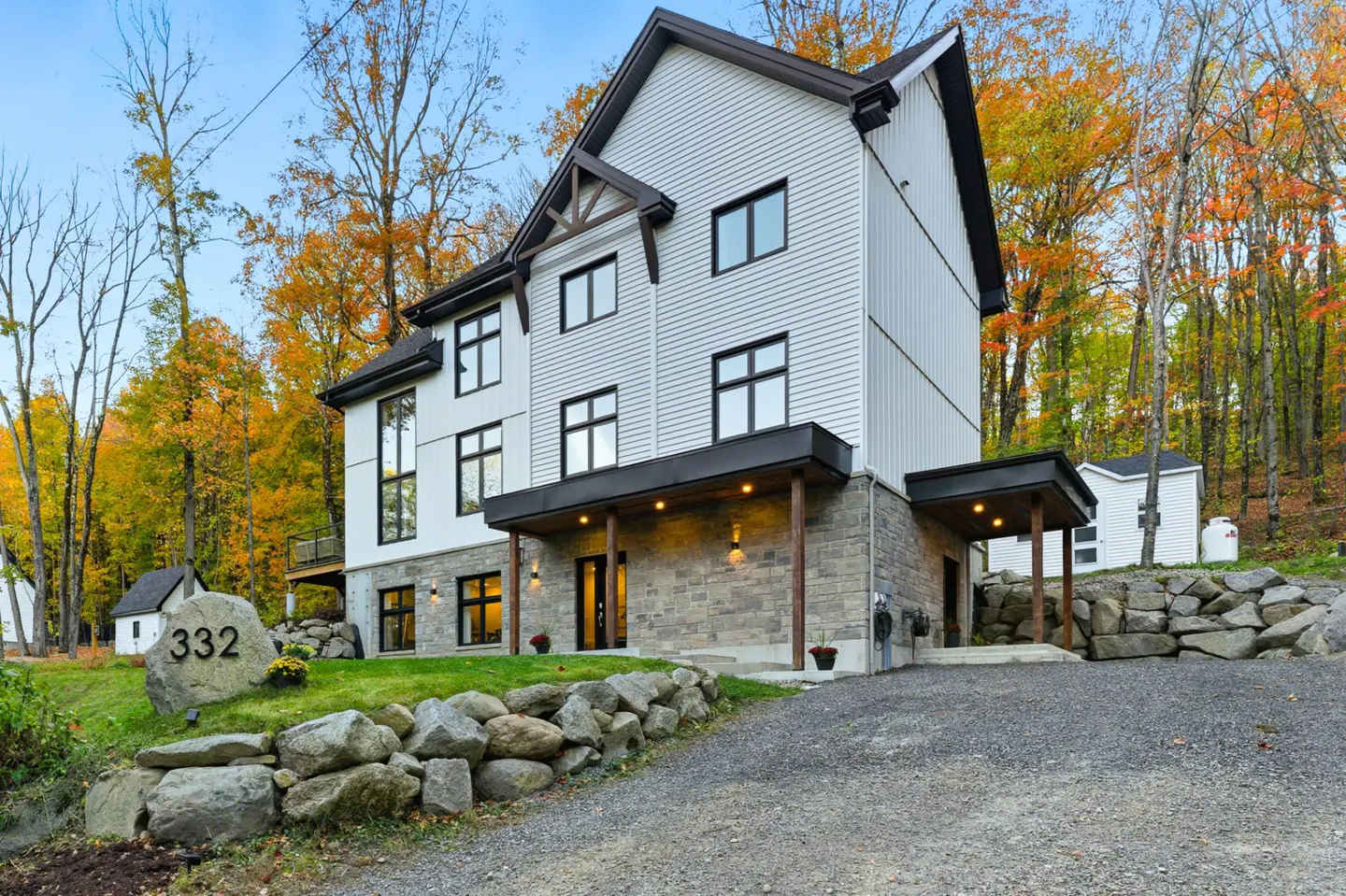 Three-story house with white siding and stone base, surrounded by trees with autumn foliage. A stone with the number 332 sits on the lawn.