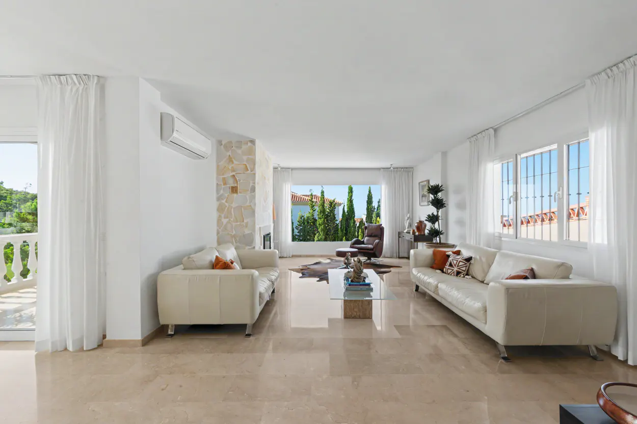 Bright living room with white sofas, marble floors, and a stone fireplace. Large windows offer views of greenery.