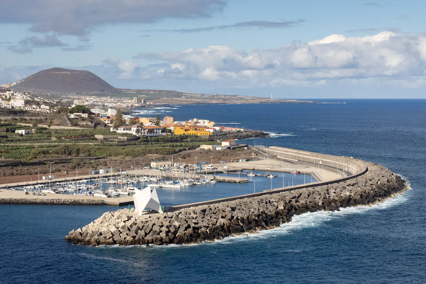 Coastal view of a marina with boats, a rock breakwater, and a white geometric structure. Buildings and a mountain are in the background.