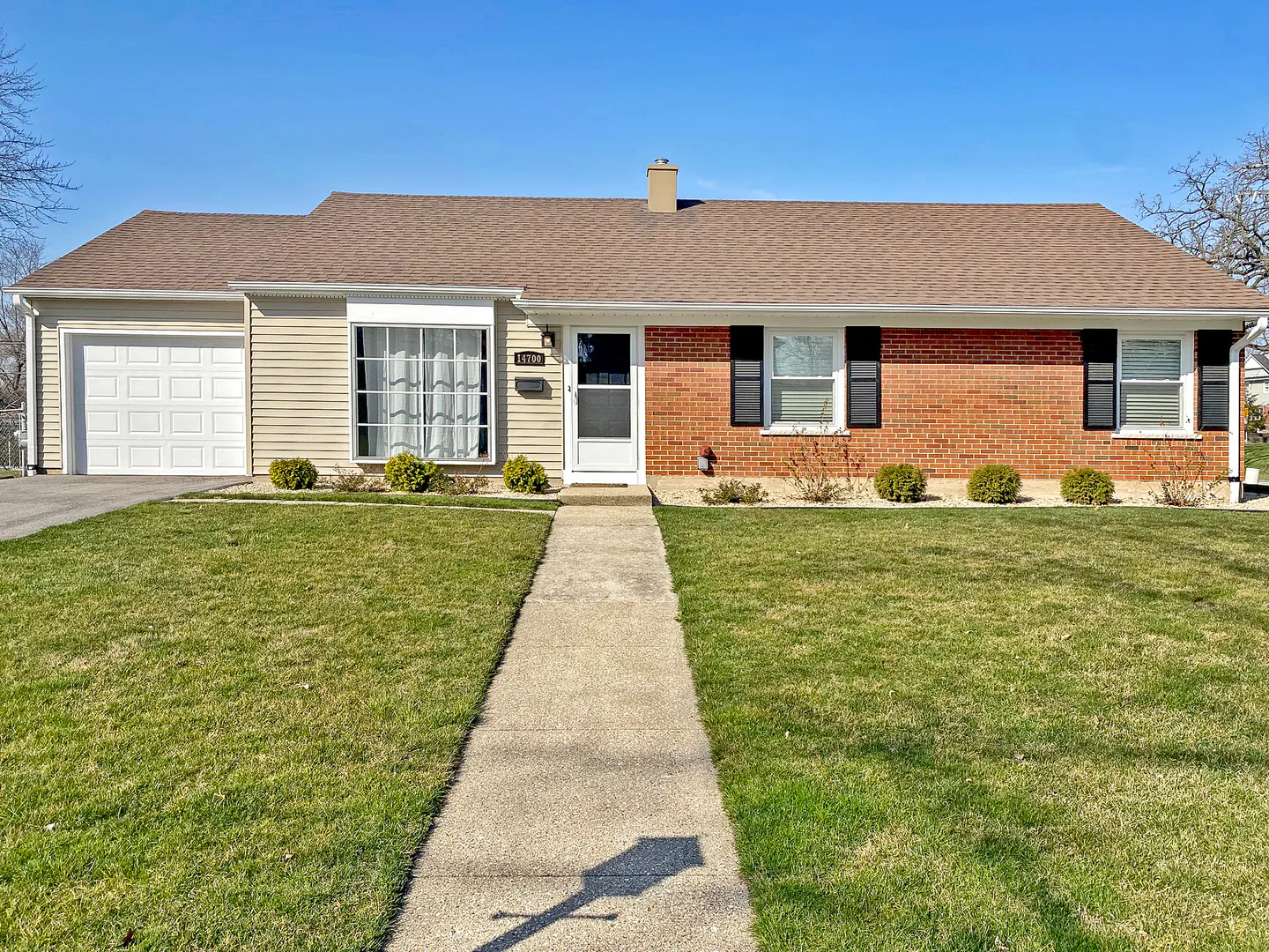 A ranch-style home with a brick and beige siding exterior, brown roof, and a green lawn on a sunny day.