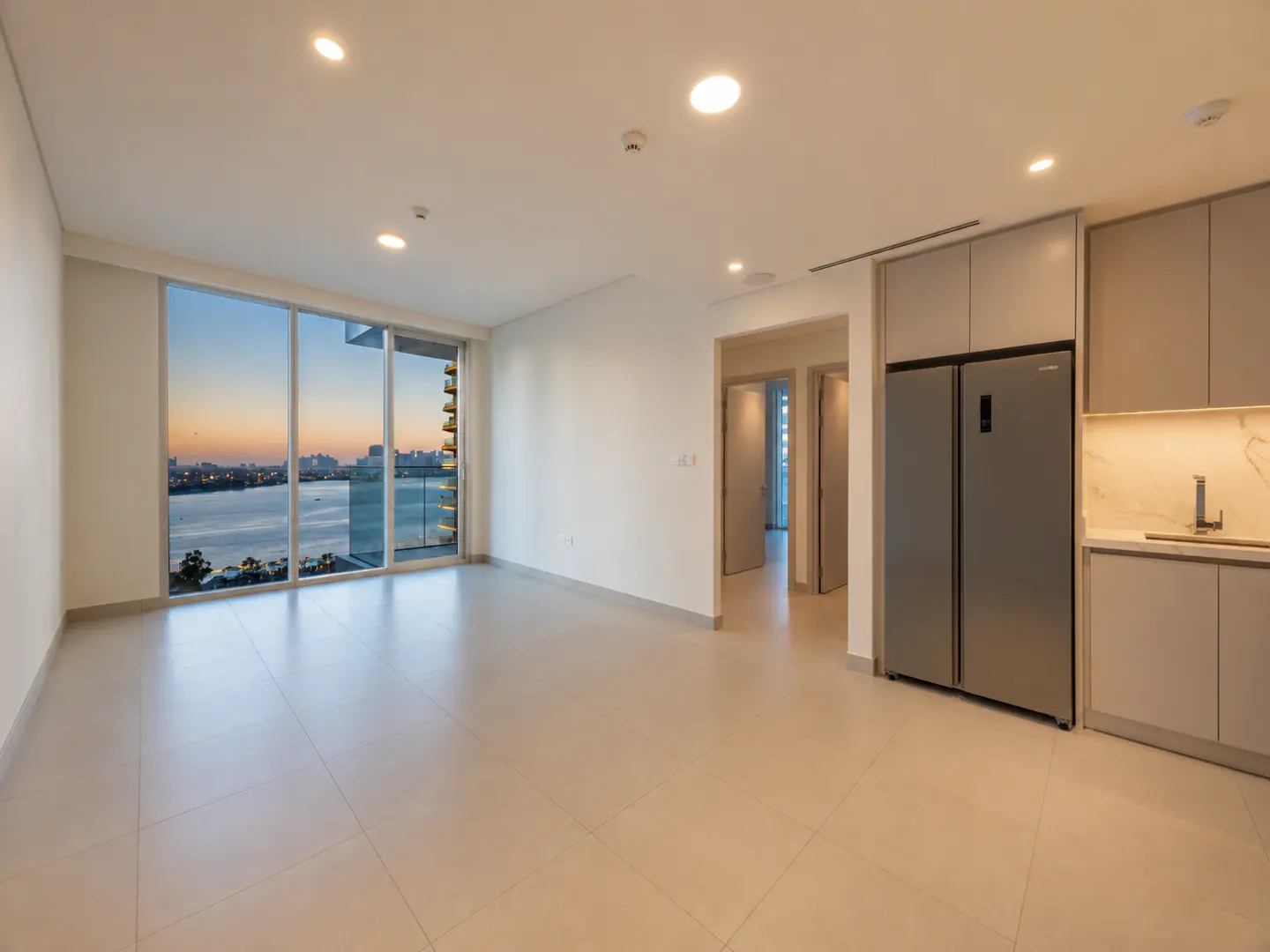 An empty, modern apartment with a large window overlooking a city skyline at sunset. The kitchen has gray cabinets and a stainless steel refrigerator.