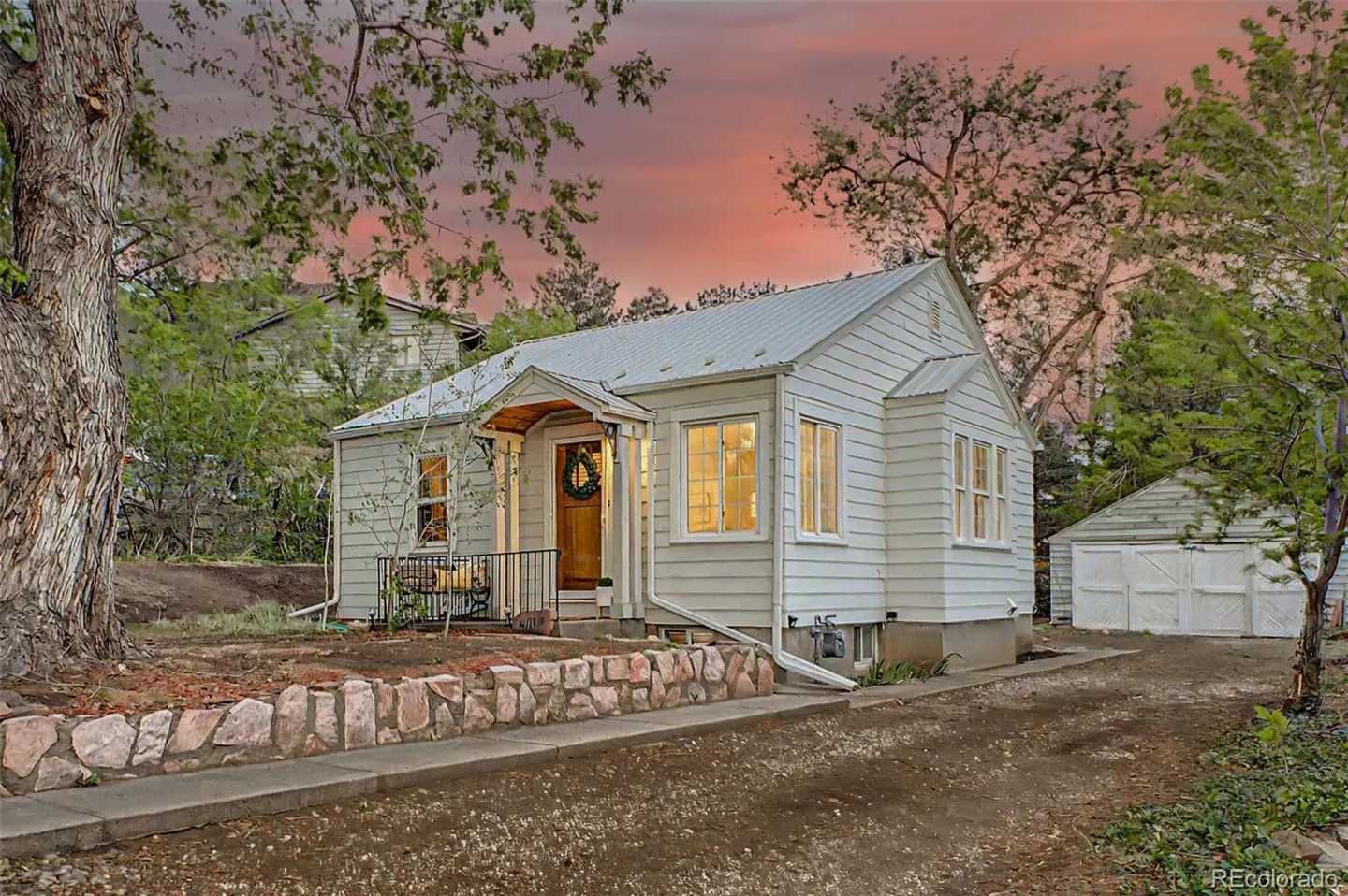 A white, one-story house with a metal roof and a small porch with a wreath on the door. A stone wall lines the front yard.