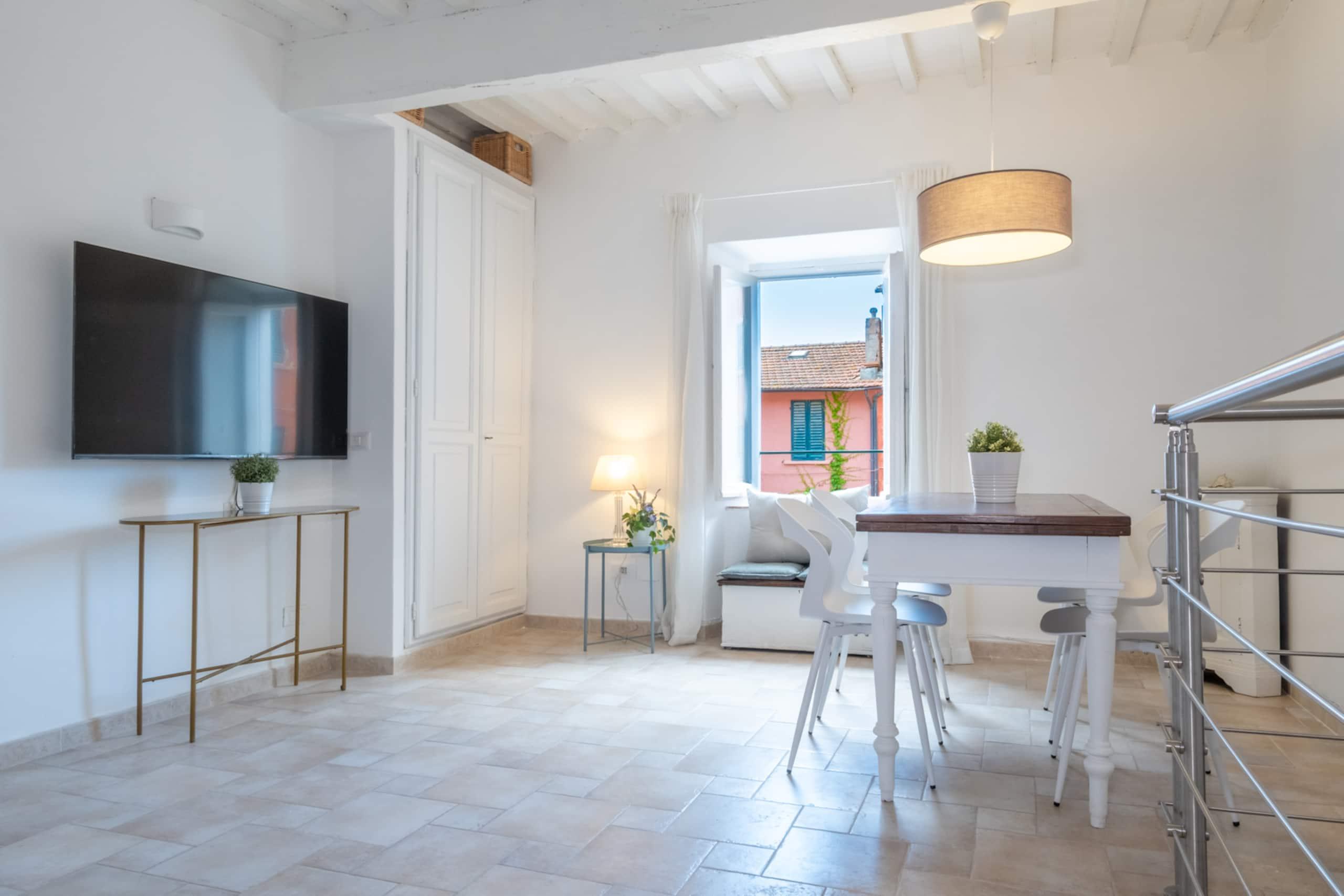 Bright dining area with white walls, tiled floor, dining table, modern chairs, wall-mounted TV, and a view of a red house. [AI-generated text]