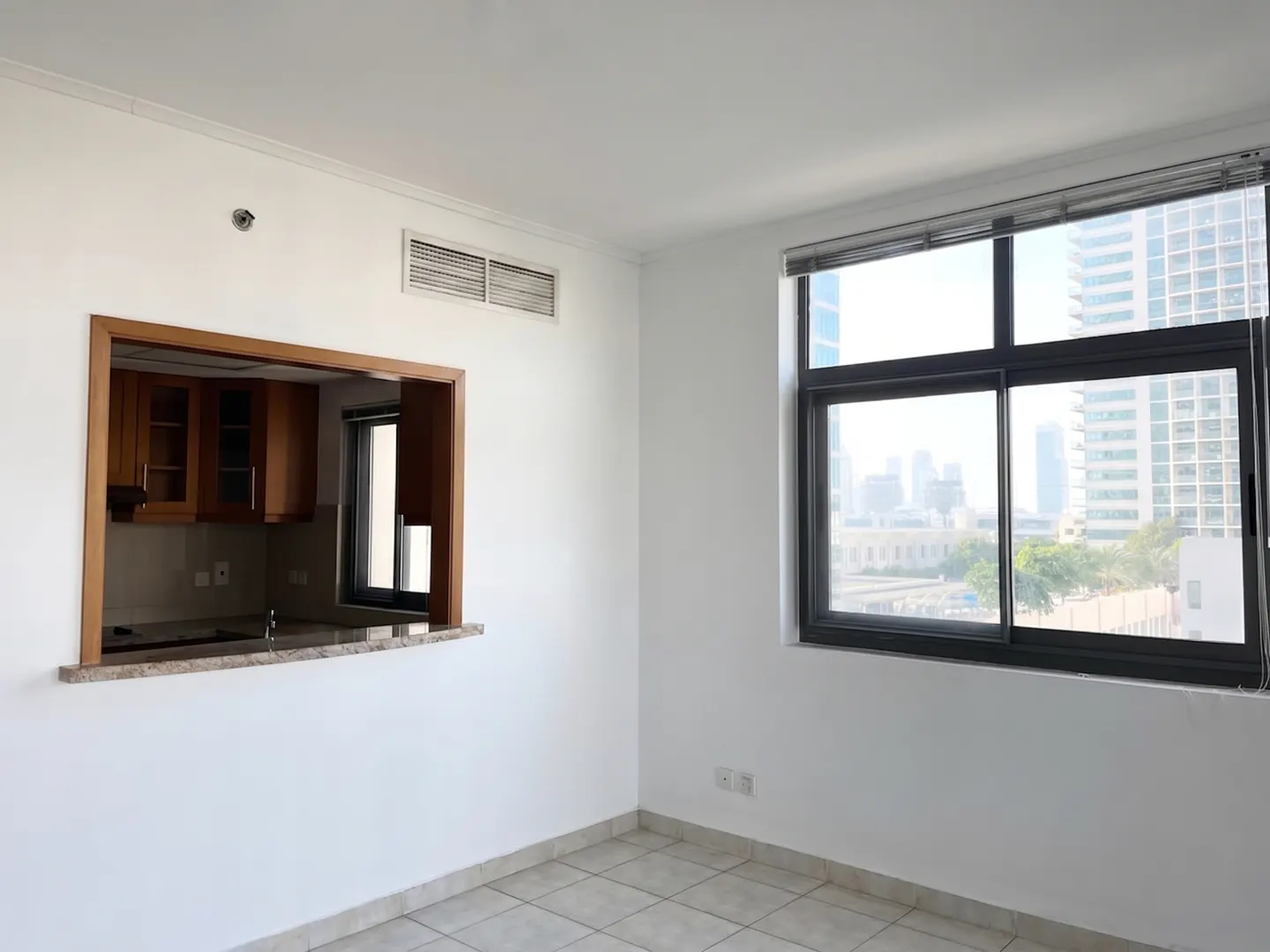 Bright, empty room with white walls, tiled floor, and a window with a city view. A kitchen pass-through with wood cabinets is visible.