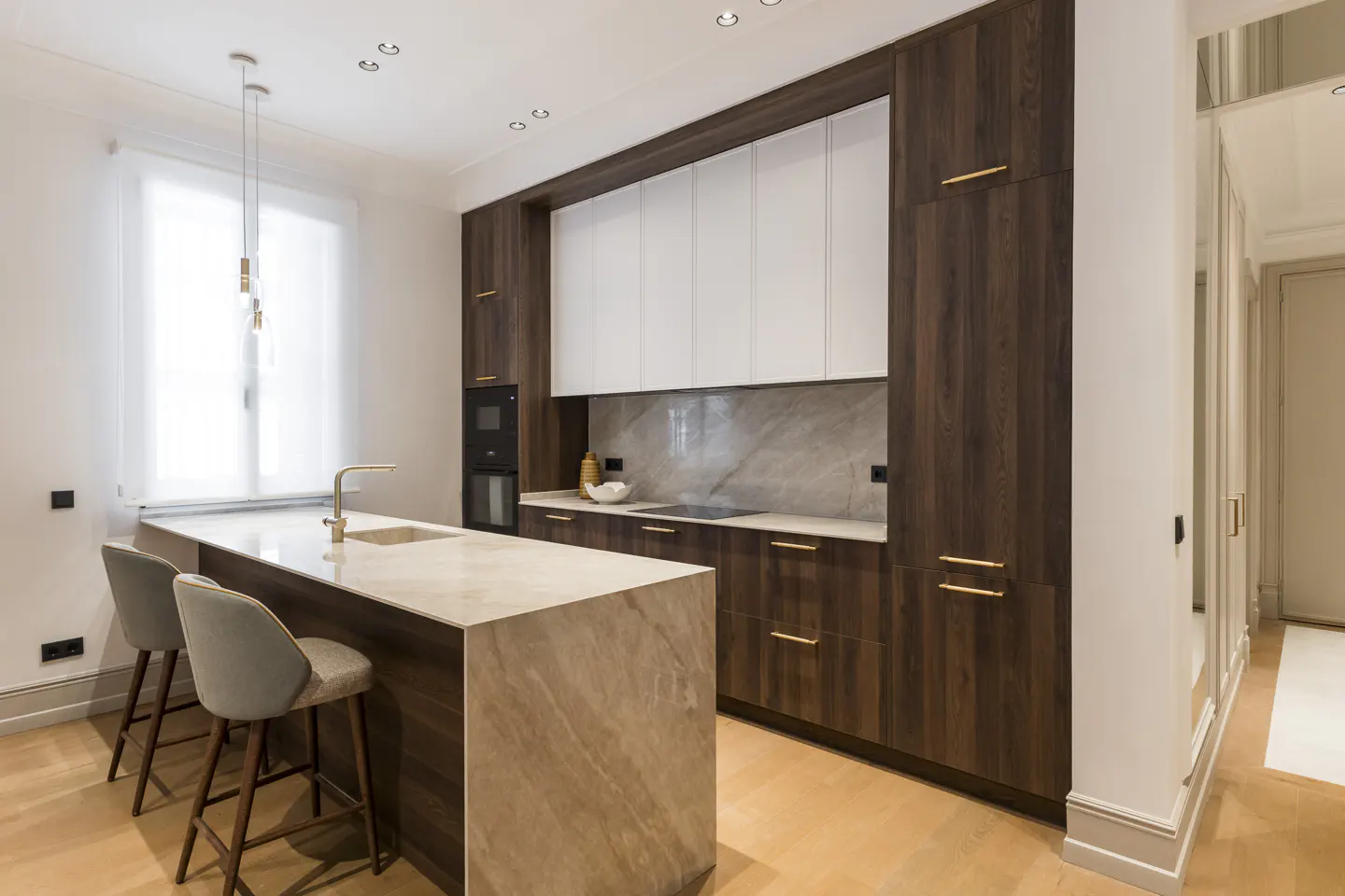 Modern kitchen with wood cabinets, white upper cabinets, and a marble island with two gray stools. A gold faucet is on the island.