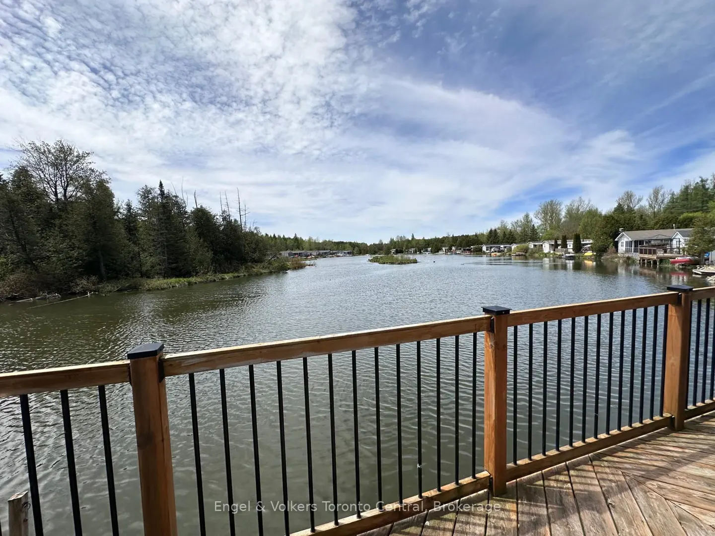View from a wood deck with black railings overlooking a calm lake surrounded by trees and houses under a cloudy blue sky.