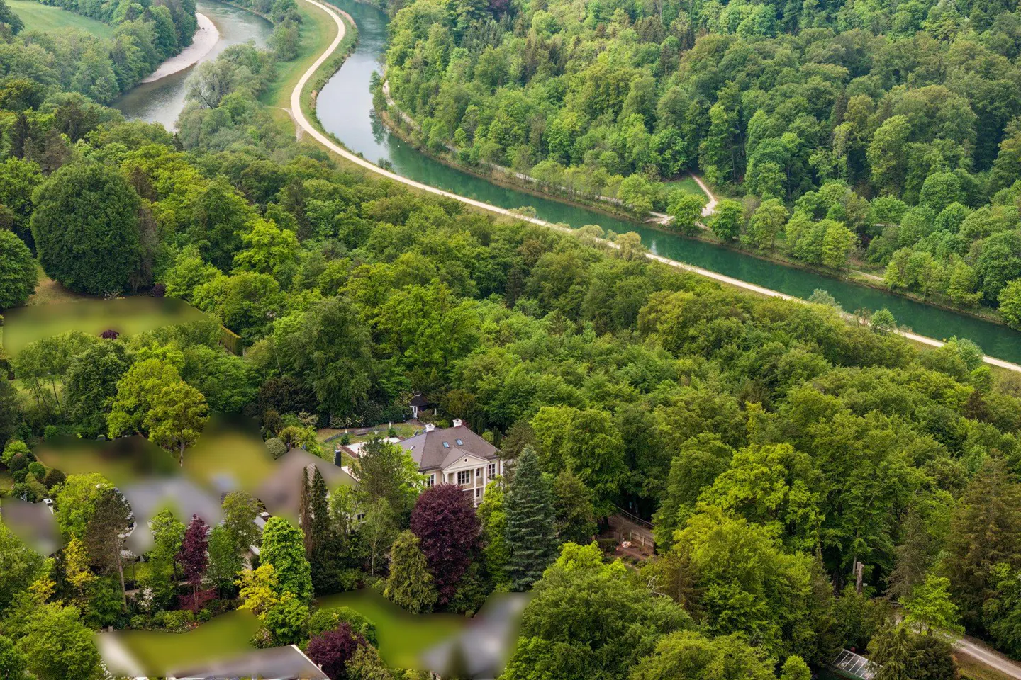 Aerial view of a house nestled in a lush green forest next to a winding river with a walking path along the bank.