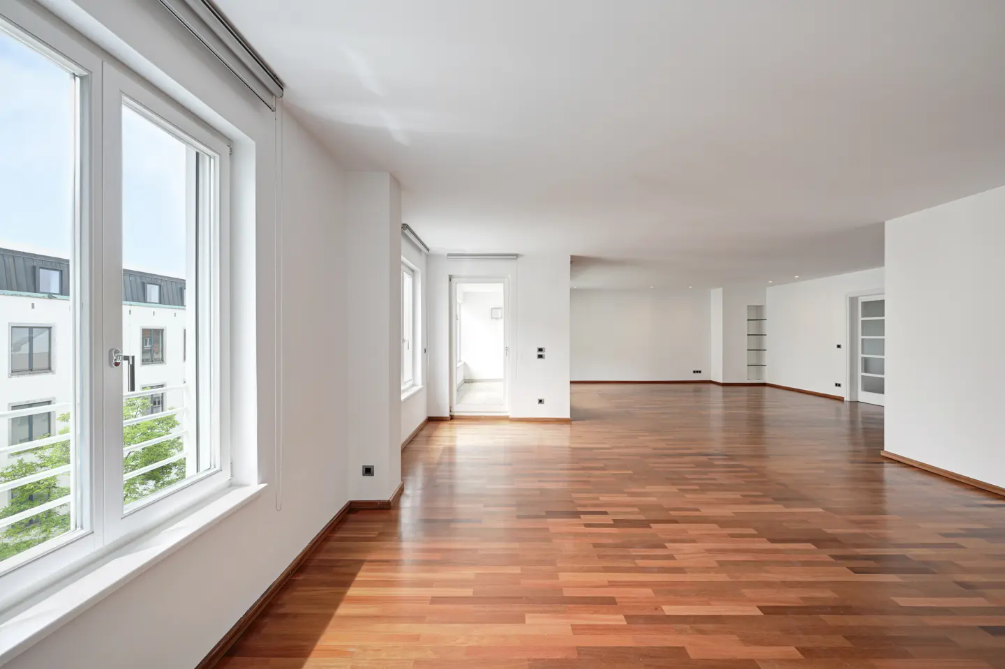 Spacious, empty room with white walls, polished hardwood floors, and large windows overlooking a neighboring building.