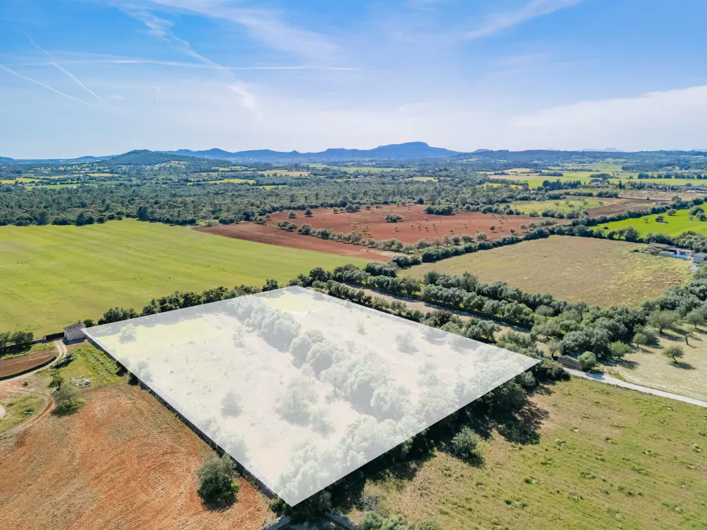 Aerial view of a large, undeveloped plot of land, outlined in white, surrounded by green and brown fields under a blue sky.