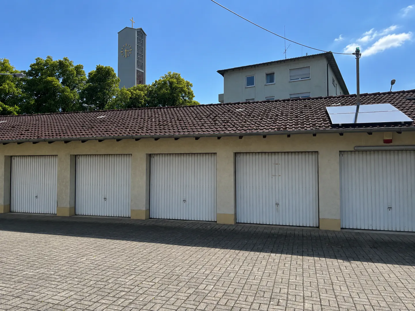 A row of five white garage doors with a brown tiled roof, solar panels, and a church tower in the background.