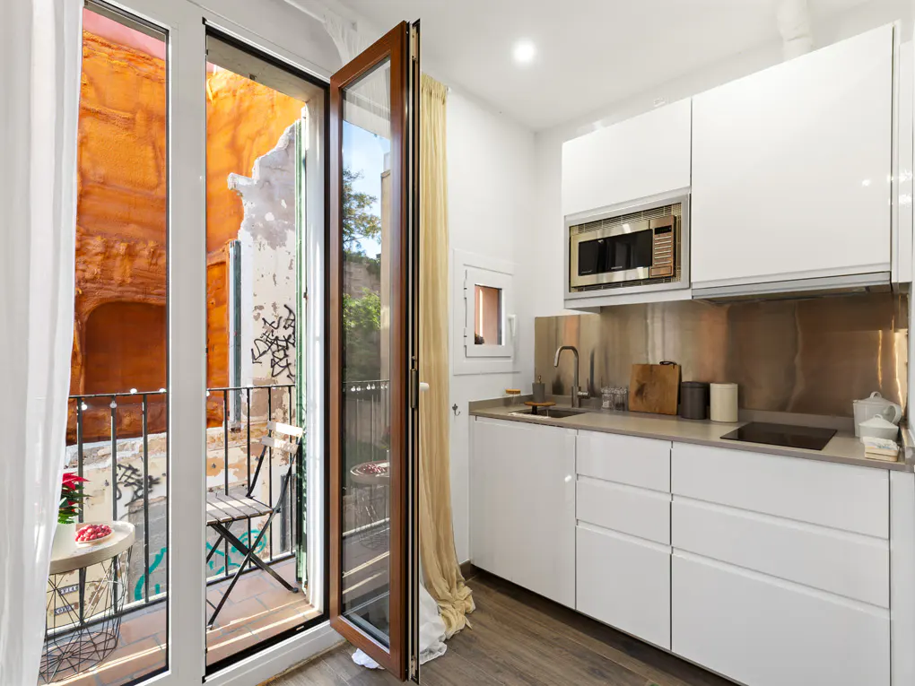 A modern white kitchen with stainless steel backsplash and open doors to a balcony with a table and chairs.