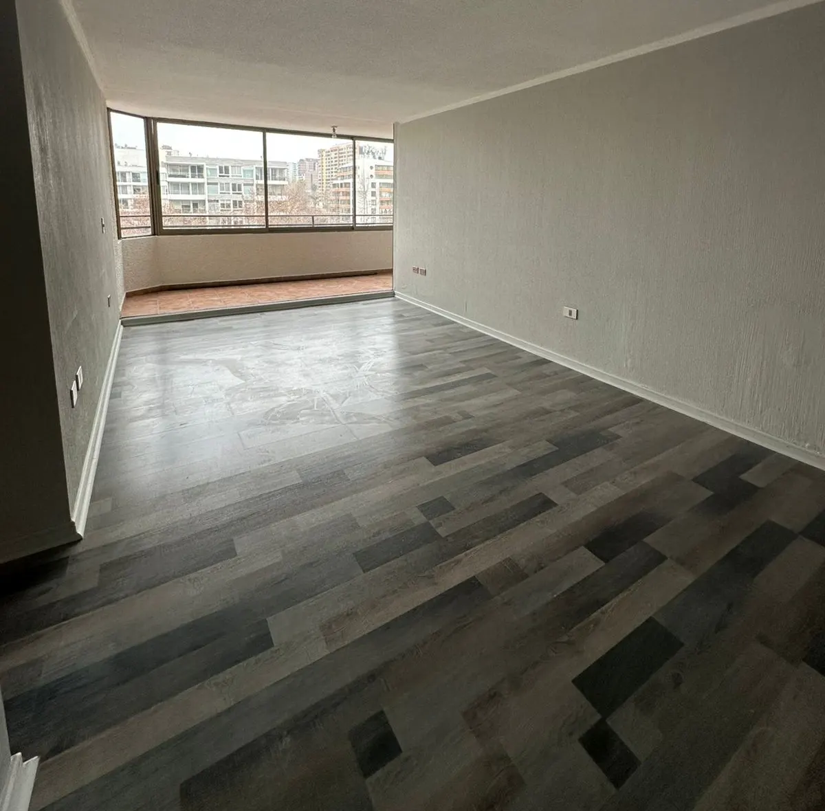 Empty living room with grey wood-look flooring, white walls, and a large window overlooking a city balcony.