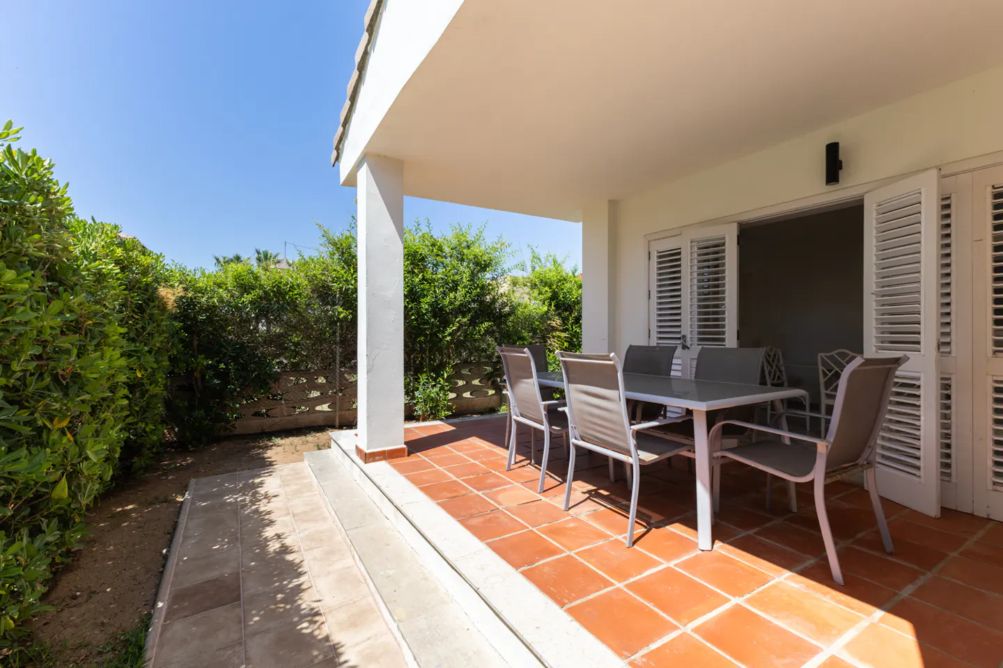 A covered patio with terracotta tiles, a dining table, and four chairs, set against a lush green hedge under a blue sky.