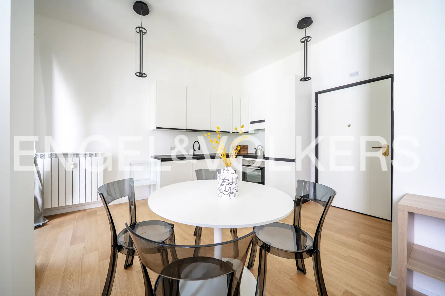 Modern kitchen with white cabinets, a round white table, and four transparent grey chairs on light wood flooring.
