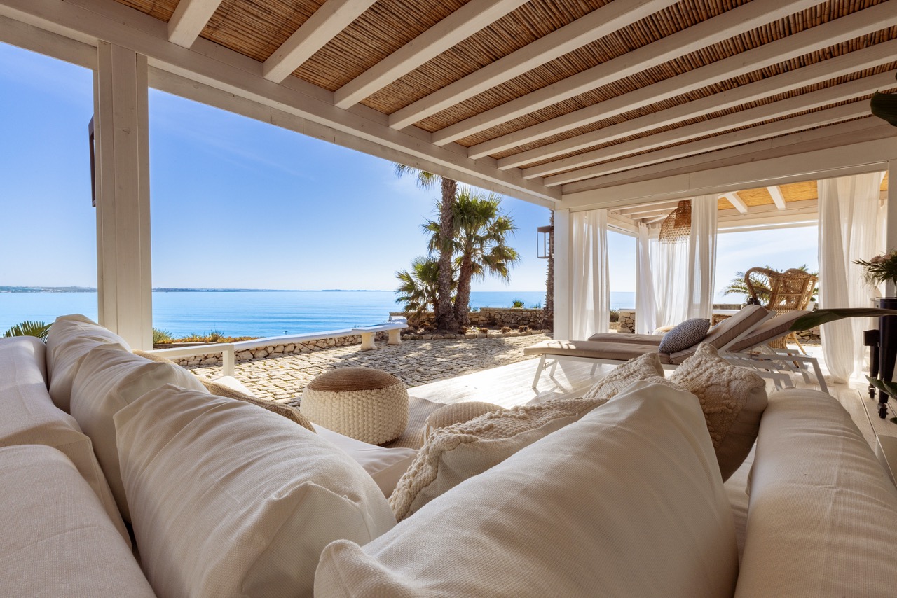 Outdoor patio with white sofa, pillows, and ottoman. Ocean view with palm trees and blue sky. White pergola with bamboo ceiling and white curtains.