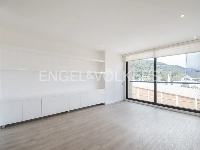 Bright, minimalist room with white built-in shelving, light wood floors, and large windows overlooking a green hillside.