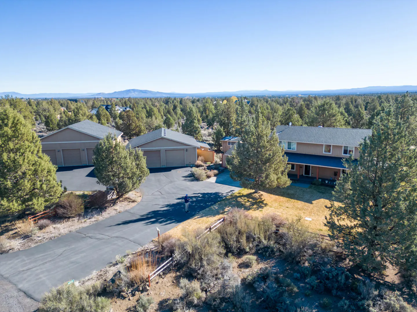 Aerial view of a tan two-story house with a blue porch roof, two garages, and a driveway surrounded by green trees.