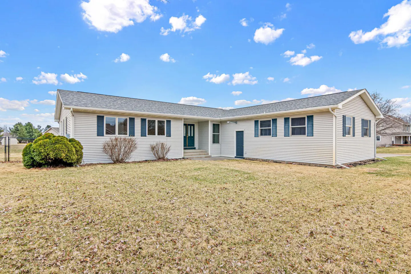 A one-story white house with blue shutters and a green door sits on a brown lawn under a blue sky.
