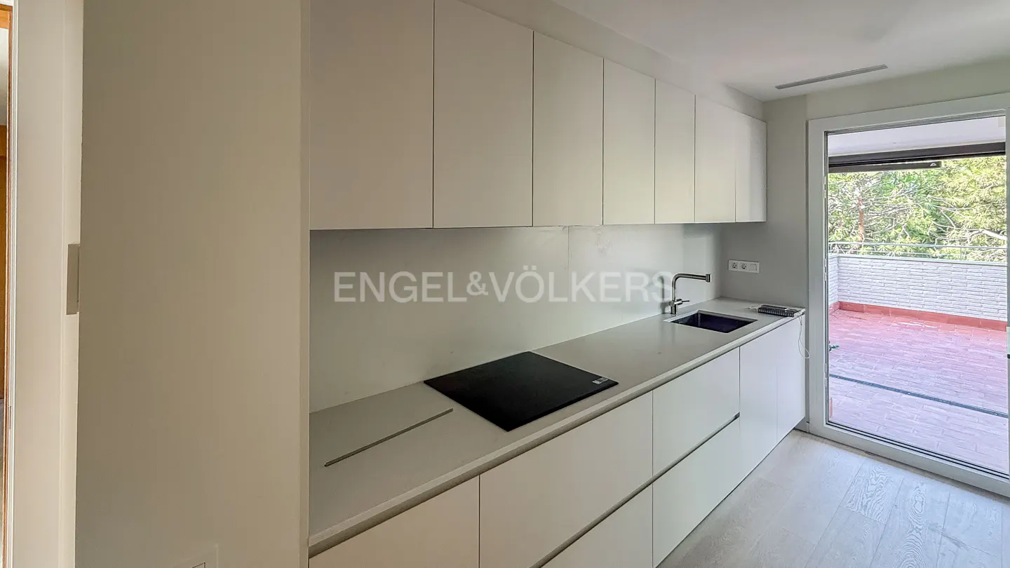 A modern, all-white kitchen with sleek cabinets, a black cooktop, and a sink. A sliding glass door opens to a brick patio with trees in the background. Engel & Völkers logo on the backsplash.