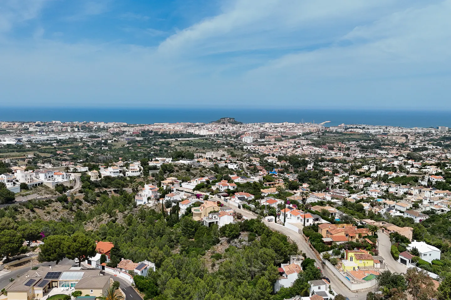 Aerial view of a hillside residential area with white houses, lush green trees, and a distant coastal town by the sea.