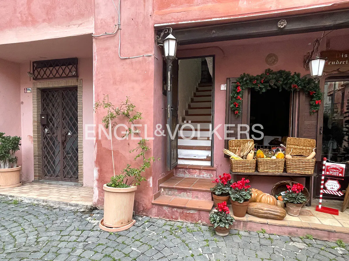 A pink building entrance with a staircase, potted plants, and baskets of produce on a cobblestone street.