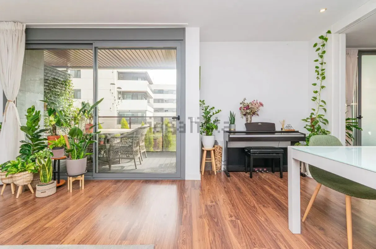 Bright living room with wood floors, a piano, and a glass door to a plant-filled balcony with outdoor seating.
