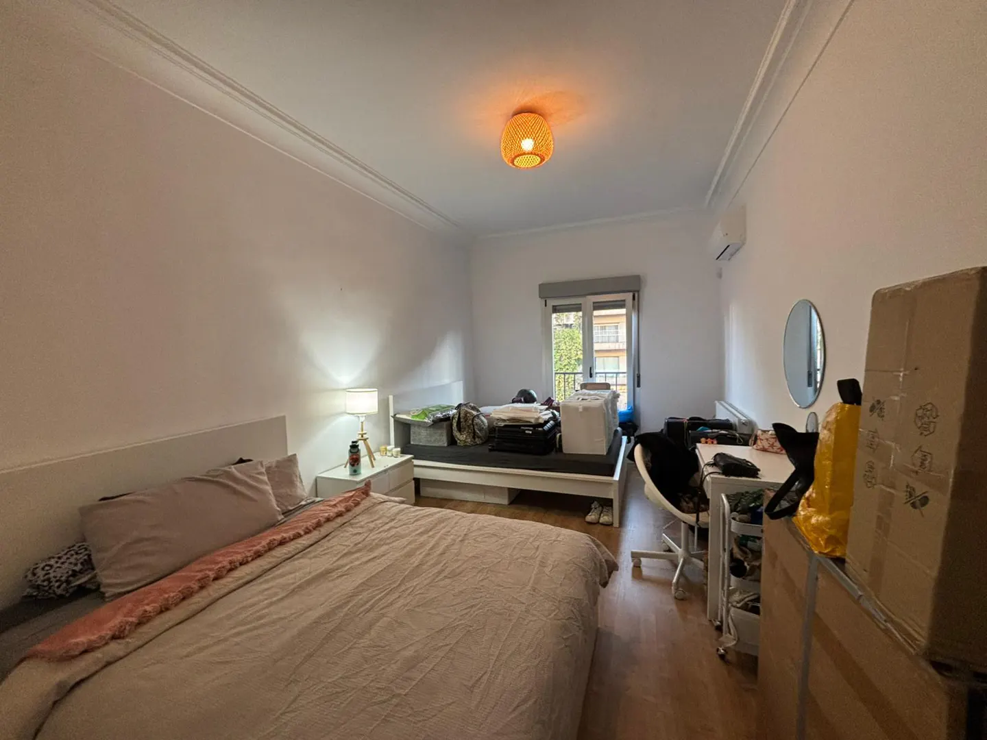 Bedroom with a large bed, a desk with a chair, a second bed with storage, and a balcony door under a woven ceiling light.