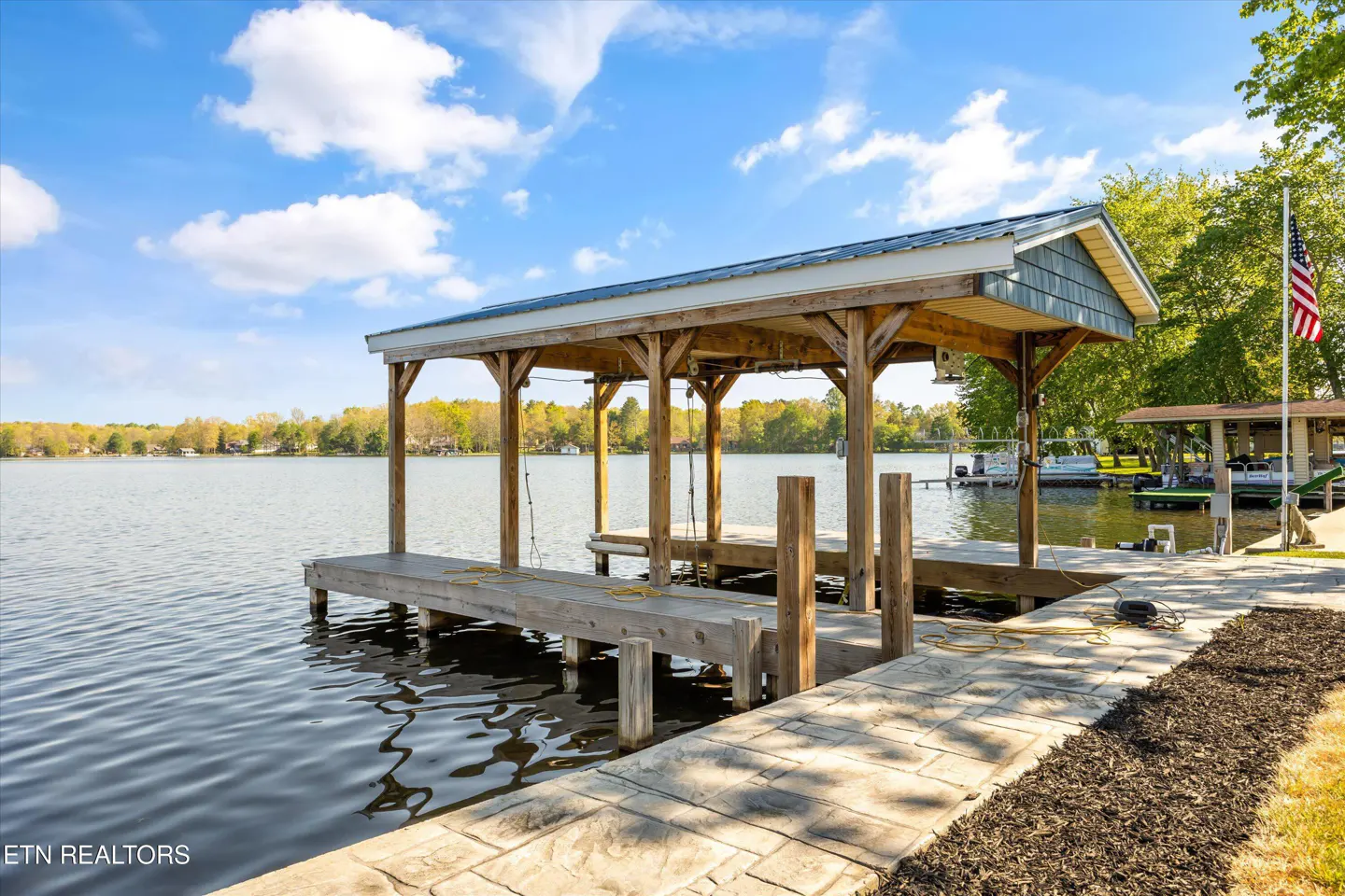 A lakeside property featuring a wooden boat dock with a covered slip, a stone walkway, and an American flag.