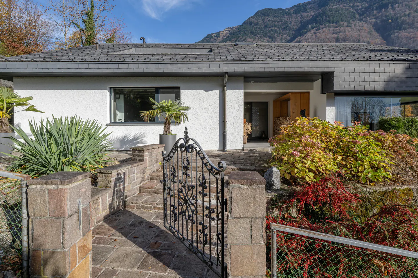 Exterior view of a white house with a gray roof, stone walkway, and black iron gate. Mountains are visible in the background.