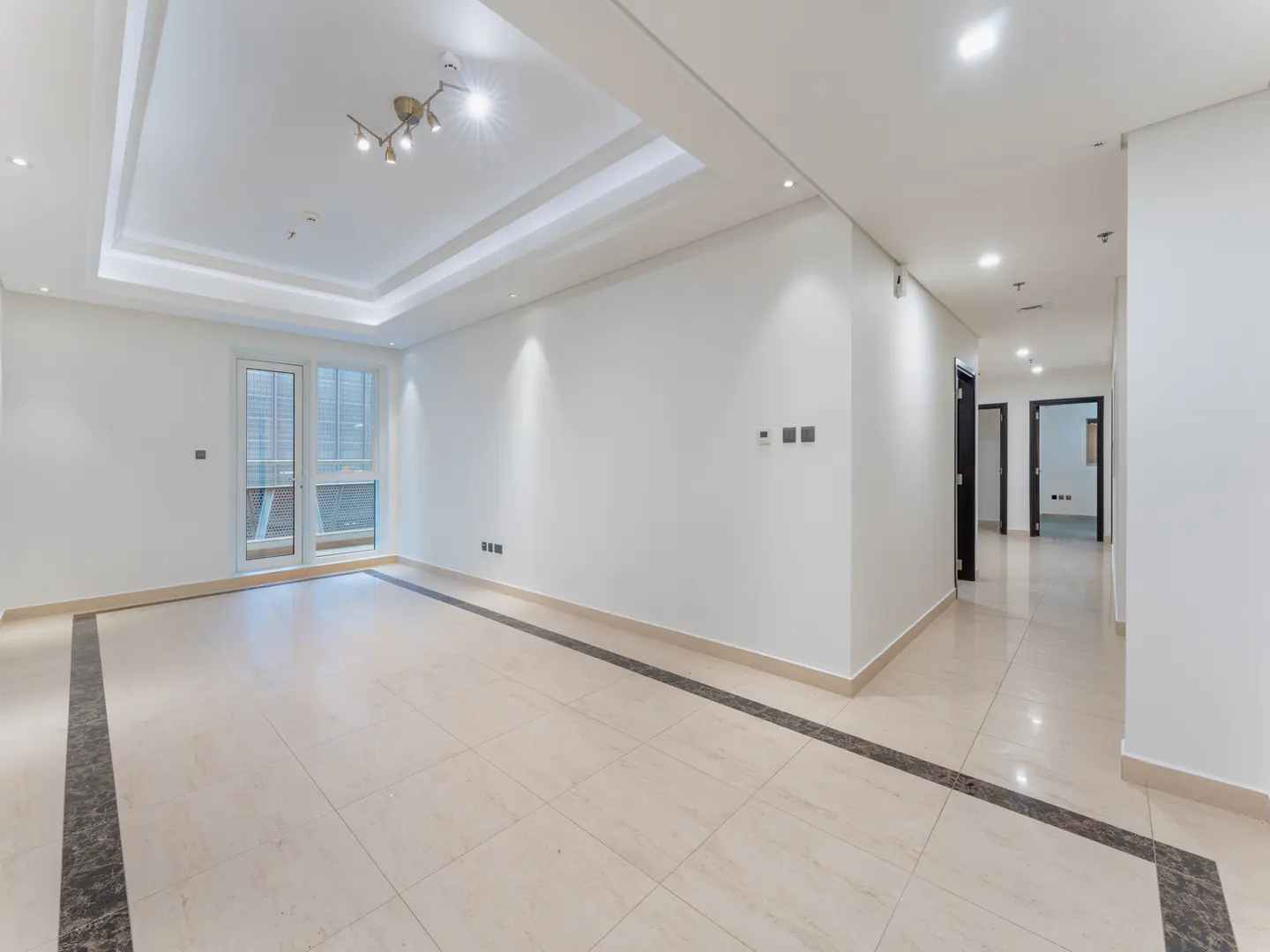 Bright, empty room with white walls, beige tile floor with dark border, and a hallway with dark doors. A window and modern light fixture are visible.