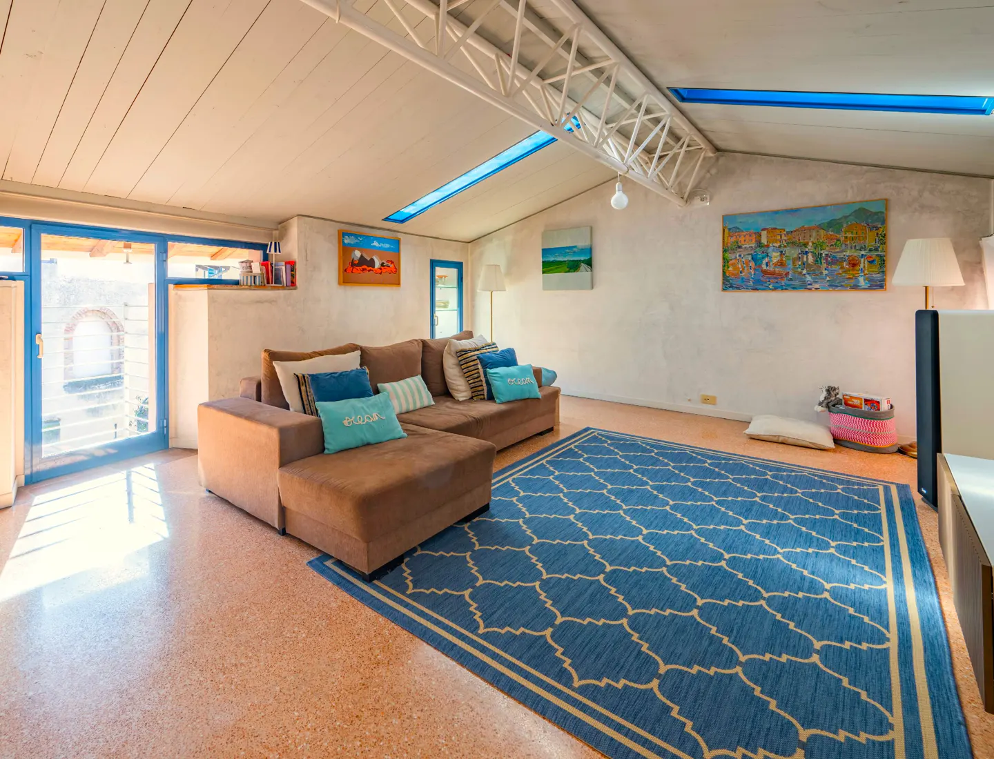 Living room with brown sectional sofa, blue patterned rug, and blue-framed sliding glass doors. Art adorns the white walls.