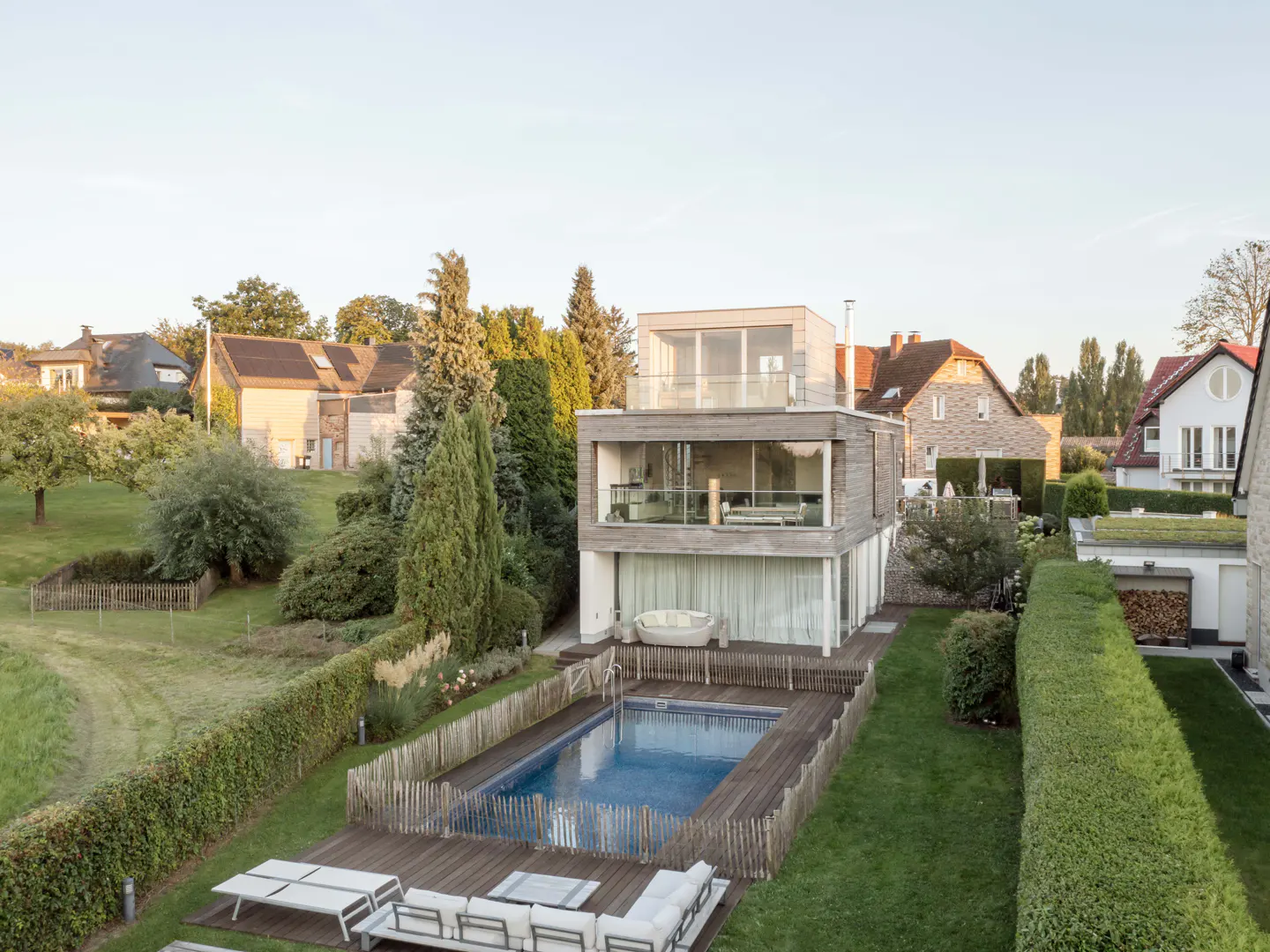 Aerial view of a modern two-story house with a pool, wooden deck, and white lounge chairs in a green backyard.