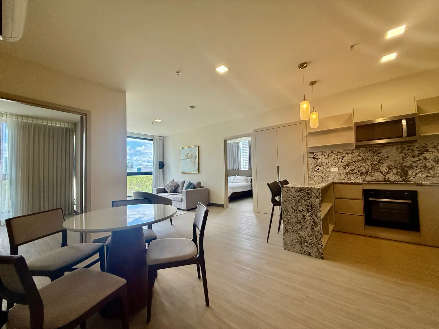 A bright, modern apartment interior with a dining table, sofa, kitchen, and bedroom visible.